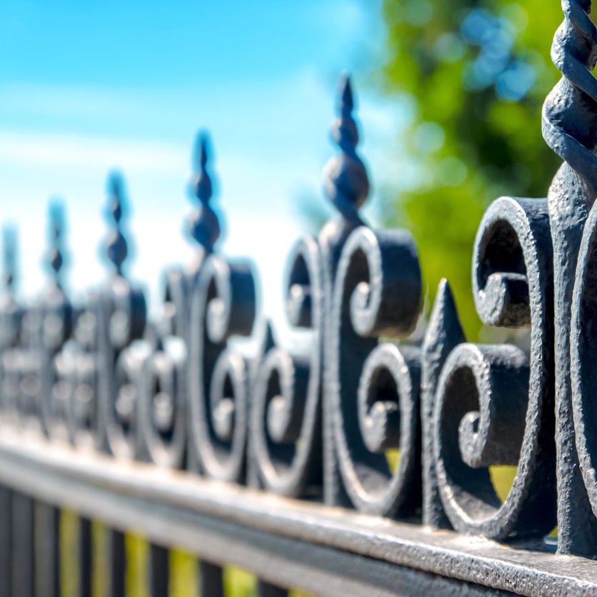 Horizontal composition photography of close-up in selective focus of iron fence, barrier, ornate with volute and curve. The black railing is taken in dimishing perspective on blurred blue sky and green tree background in sunny day of summer season.